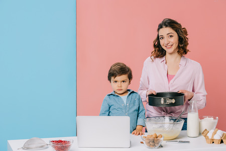 mother holding baking form while cooking together with little son at kitchen table with products,...