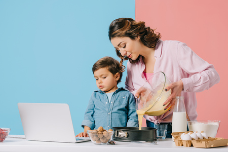mother pouring dough into baking form while little son looking at laptop display on bicolor backgroundの写真素材