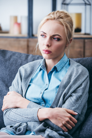 selective focus of beautiful upset woman with arms crossed sitting on couch at homeの写真素材