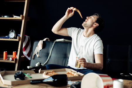 man eating delicious pizza while holding bottle in messy living roomの写真素材