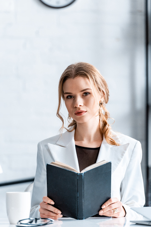 beautiful businesswoman sitting at desk, holding notebook and looking at camera at workplaceの写真素材