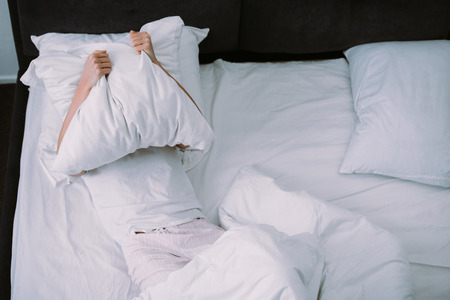 depressed woman covering face with pillow while lying in bed at home