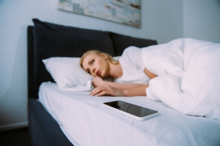 upset woman lying with smartphone in bed at home