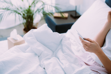 cropped view of woman using smartphone in bed at homeの写真素材