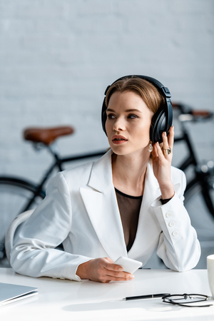 selective focus of businesswoman in headphones sitting at desk and using smartphone at workplaceの写真素材