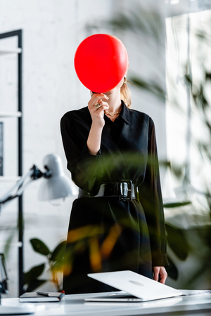 woman in black clothes standing near notebook and hiding face behind red balloonの写真素材