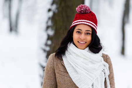 african american young woman in warm clothing looking at camera and smiling in winter park
