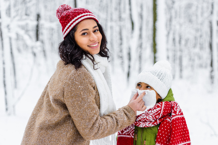 african american woman looking at camera and wiping with napkin nose of preteen daughter in winter parkの写真素材