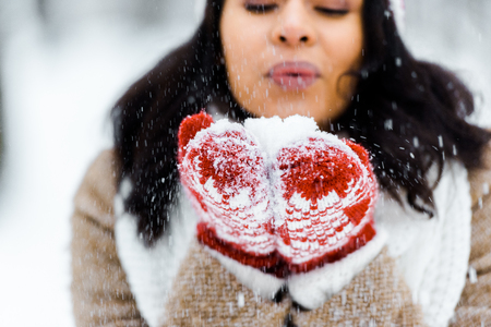 cropped view of beautiful african american woman blowing snow in winter forest and looking at snowの写真素材