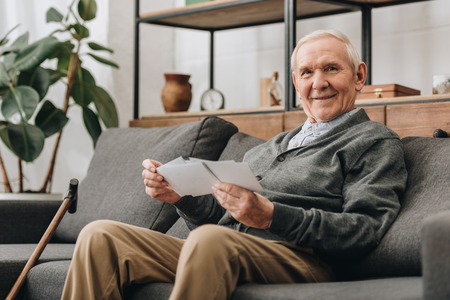 cheerful senior man with grey hair holding photos and sitting on sofaの写真素材