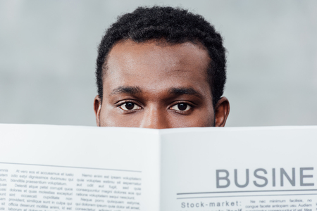 african american casual businessman holding newspaper in front of face and looking at camera