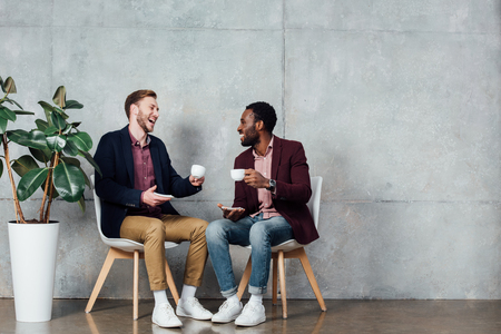 happy multiethnic men sitting, drinking coffee and talking in waiting hallの写真素材