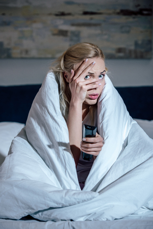 frightened woman covering face with hand while watching tv in bed at homeの写真素材