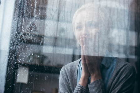 sad adult woman with praying gesture crying at home through window with raindropsの写真素材