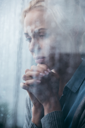 sad adult woman with folded hands at home through window with raindrops