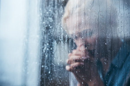 sad adult woman with folded hands at home through window with raindropsの写真素材