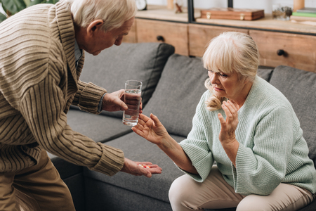 senior man looking at old woman and giving glass of water with pillsの写真素材