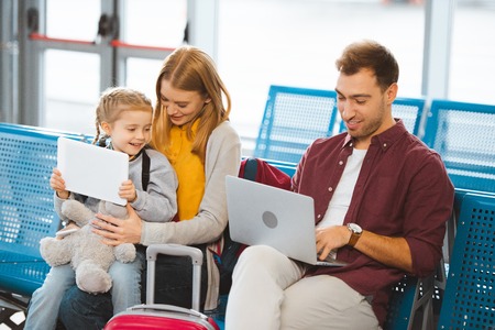 cute kid holding digital tablet near mother and looking at father using laptop in airportの写真素材