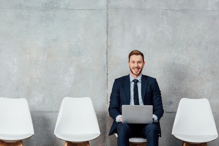 smiling businessman sitting on chair and using laptop in waiting hall