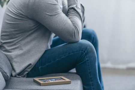 cropped view of man in grey shirt and blue pants sitting on couch with photo frame at home, grieving...