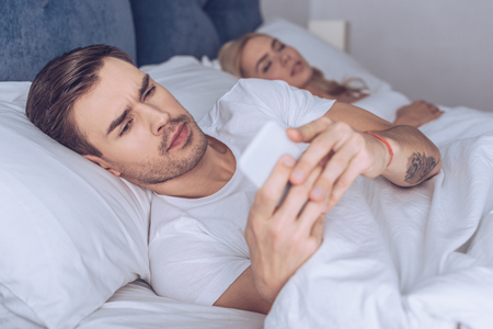 suspicious young man using smartphone while lying with sleeping girlfriend in bedの写真素材