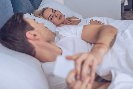 suspicious young man using smartphone and looking at sleeping girlfriend in bed, secret conceptの写真素材