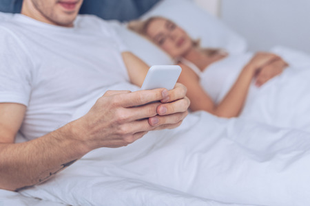 cropped shot of young man using smartphone while lying with sleeping wife in bedの写真素材