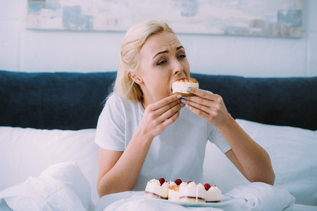 depressed woman in pajamas eating sweet cake in bed aloneの写真素材
