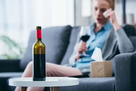selective focus of wine bottle with woman holding glass of wine and wiping tears at home on backgroundの写真素材