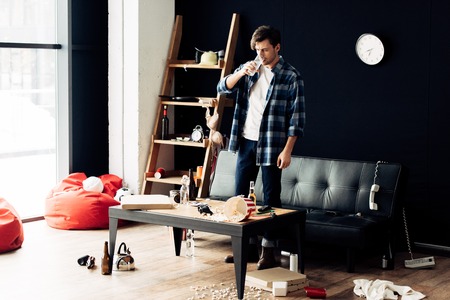 drunk man holding glass and drinking water after party in messy living room