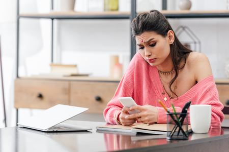 upset beautiful indian student using smartphone at table with laptopの写真素材