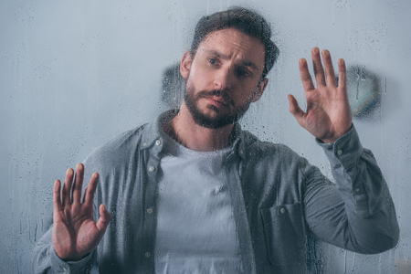 handsome upset man touching window with raindrops and looking away