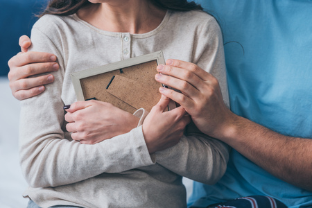 cropped view of husband hugging wife with picture frame at homeの写真素材