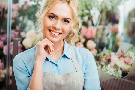 beautiful female florist in apron touching ching and smiling with flower shop on backgroundの写真素材