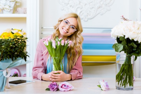 beautiful female florist looking at camera and holding vase with pink tulips in flower shop