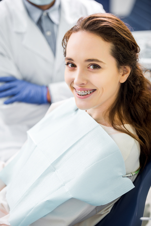 selective focus of woman in braces smiling with dentist standing with crossed arms on backgroundの写真素材