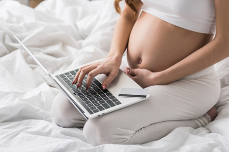 Partial view of pregnant woman sitting on bed with laptop and credit cardの写真素材