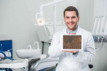cheerful dentist holding digital tablet with graphs on screen in dental clinicの写真素材