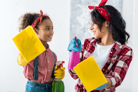 smiling african american mom and daughter with yellow rags and spray bottles cleaning houseの写真素材