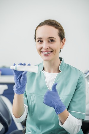 cheerful female dentist with braces on teeth holding toothpaste and showing thumb upの写真素材