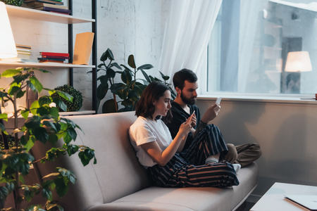 young couple sitting on couch and using smartphones in living roomの写真素材