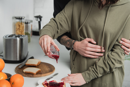 cropped view of woman pouring jam on toasts during breakfast in kitchen with man on backgroundの写真素材