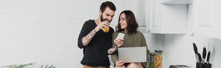 man drinking juice and using smartphone while woman using laptop during breakfast in kitchenの写真素材