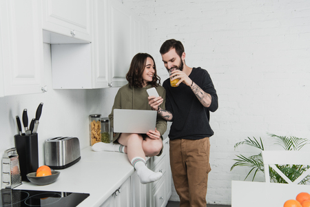 man drinking juice and using smartphone while smiling woman using laptop during breakfast in kitchenの写真素材