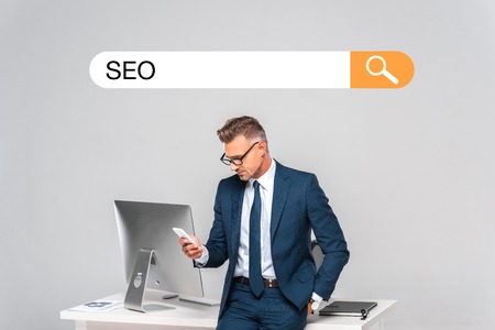 handsome businessman leaning on table and using smartphone isolated on grey with seo in search barの写真素材
