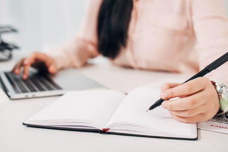 Cropped view of woman writing in notebook and typing on computer keyboard