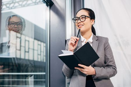 pretty businesswoman smiling, standing by window and holding notebook and penの写真素材