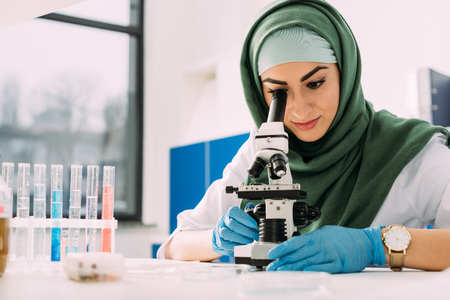beautiful female muslim scientist looking through microscope during experiment in chemical laboratoryの写真素材