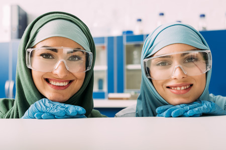 smiling female muslim chemists in protective goggles and hijab looking at camera in laboratoryの写真素材