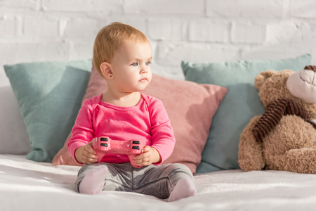 adorable kid in pink shirt holding pink joystick on bed in children room and looking awayの写真素材